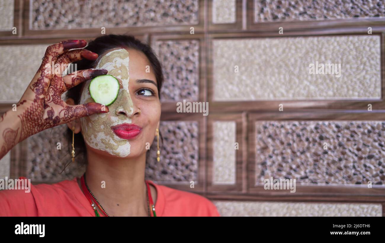 A woman with facial mask and fresh cucumber on eyes, relaxing at home. Close-up portrait of smiling girl in clay mask. homemade multani mitti face pac Stock Photo