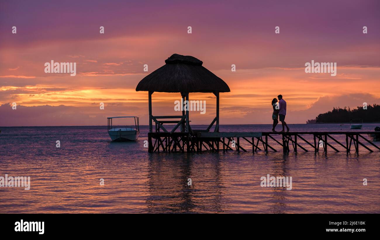 couple watching the sunset on a tropical beach with a wooden pier in ...
