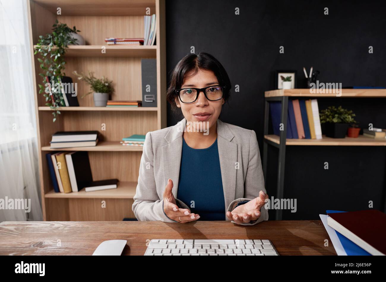 Portrait of confident Latin teacher in eyeglasses sitting in own office and gesturing hands while holding tutorial online Stock Photo