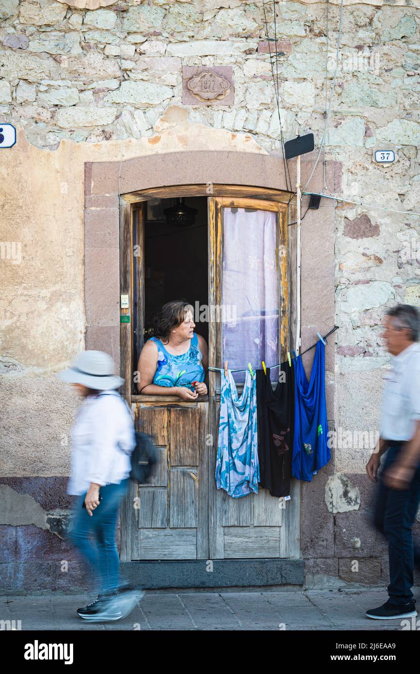 Sardinian street life - a elder woman looks out of the front door to the right onto the street while two pedestrians pass by on the pavement Stock Photo
