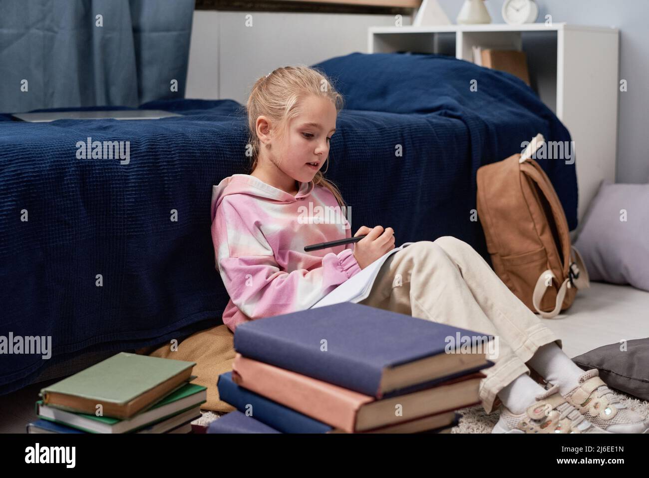 Blond-haired girl in hoodie with abstract pattern sitting at bed and reading notes while preparing for school test Stock Photo