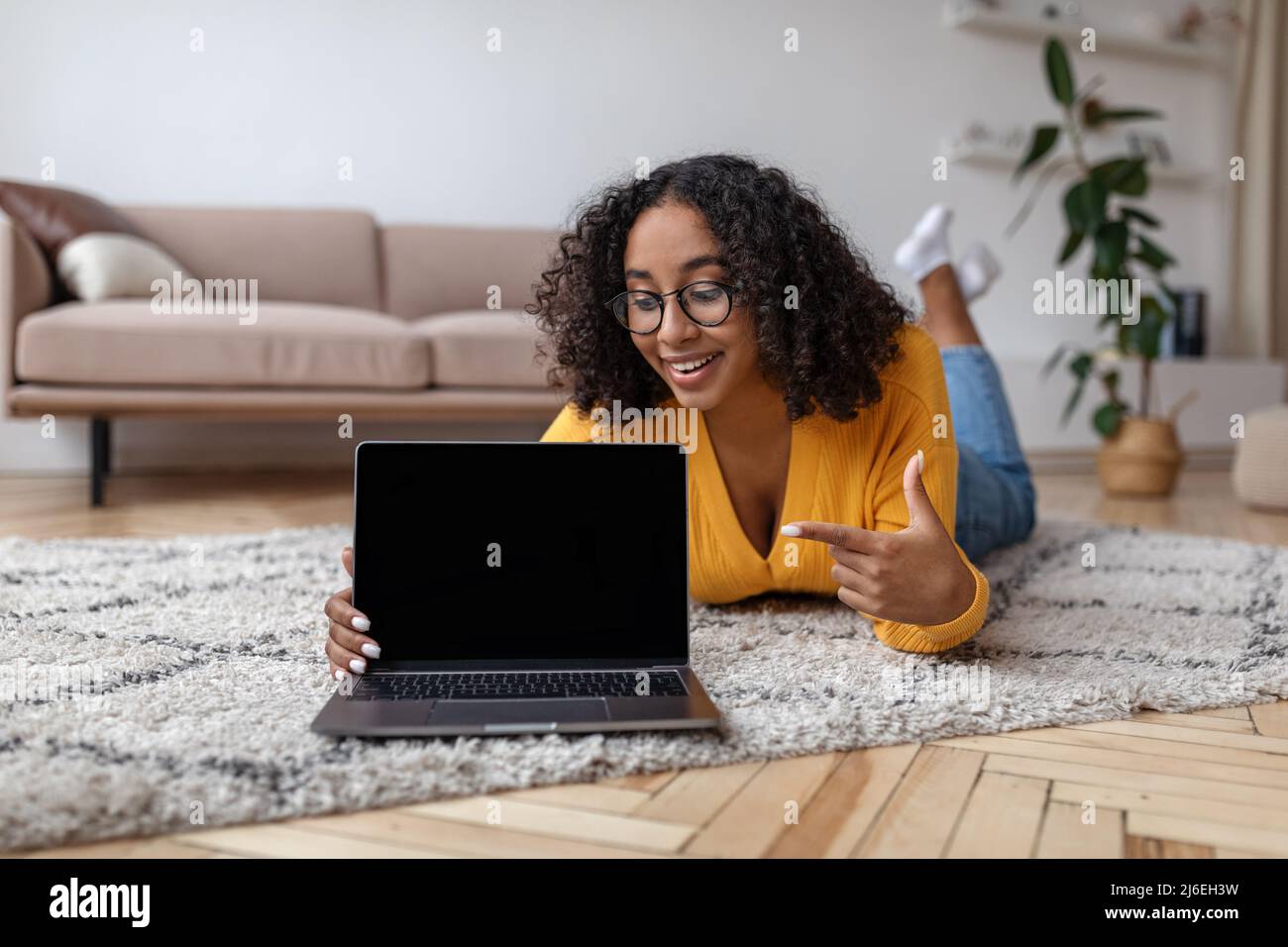 Happy young black woman lying on floor at home, pointing at laptop pc ...