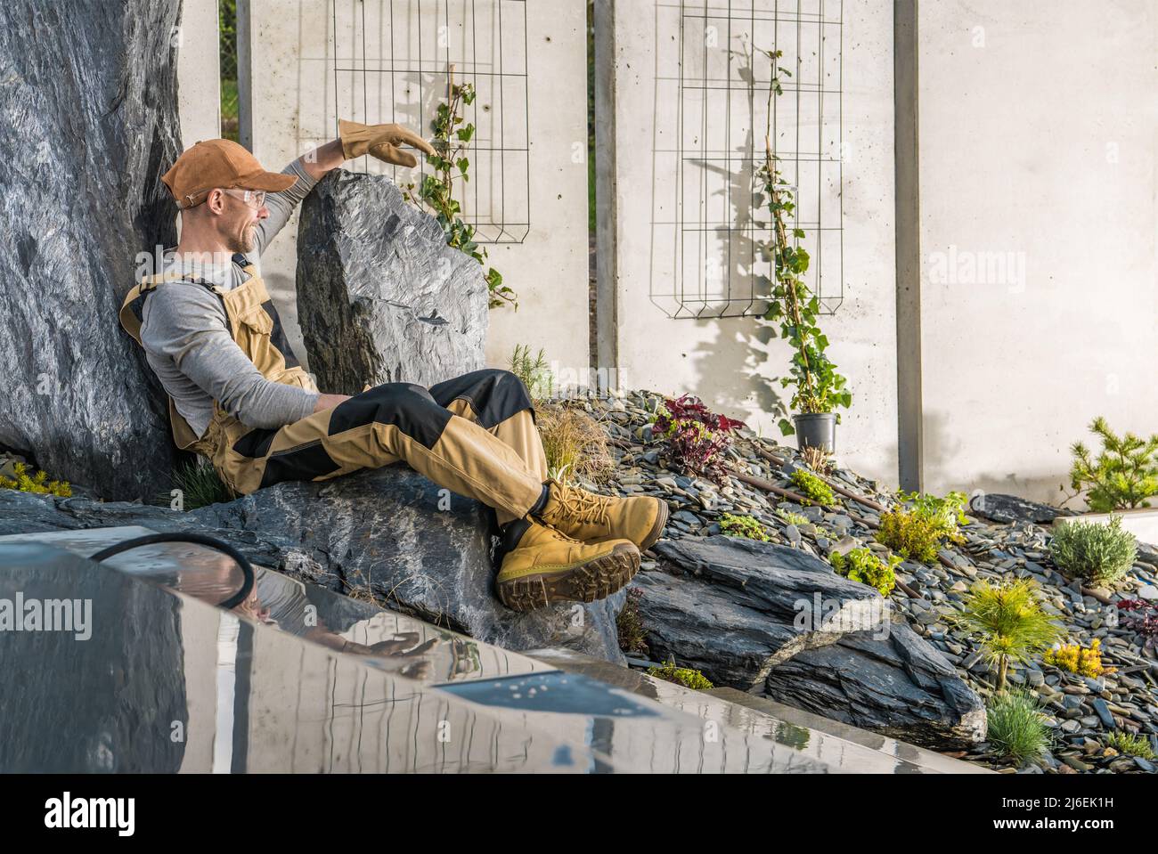 Happy Caucasian Gardener Relaxing on a Piece of Decorative Rock After Long Hard Day of Work in the Beautiful Backyard Garden. Landscaping Industry The Stock Photo