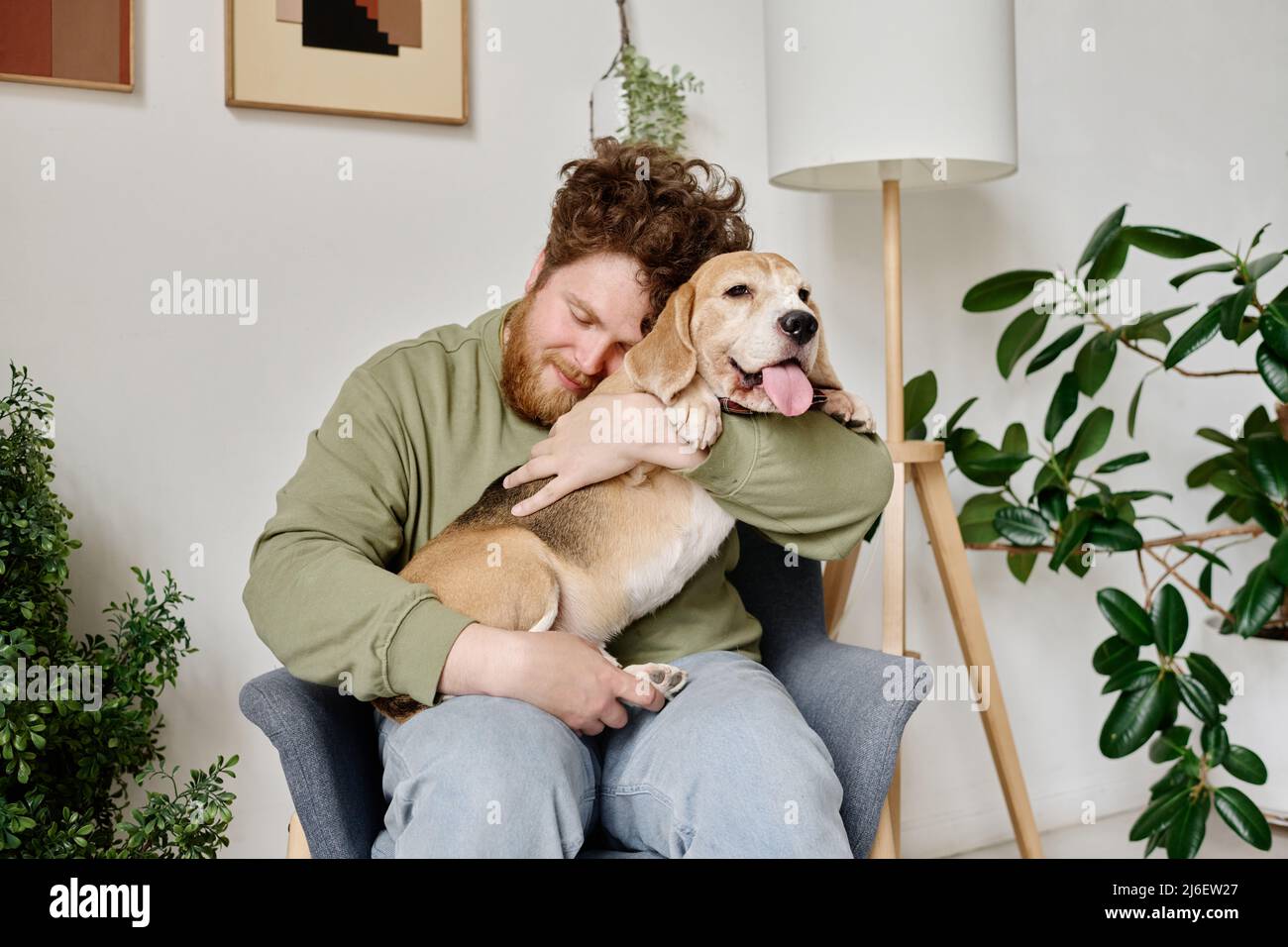 Young bearded man sitting on armchair in room with plants embracing his dog and relaxing Stock Photo