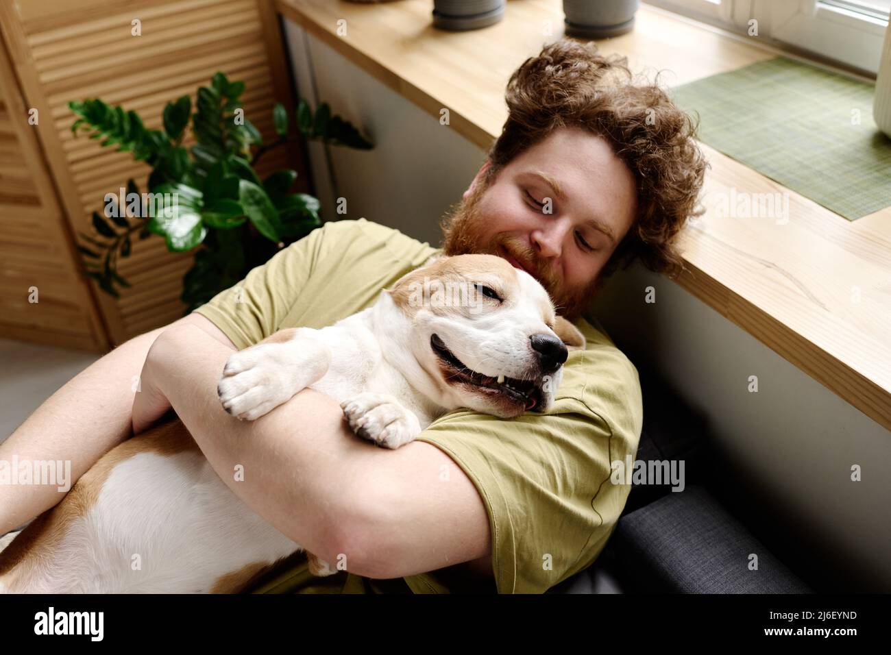 Young bearded man lying on bed and holding his sleeping dog on arms during rest time in bedroom Stock Photo