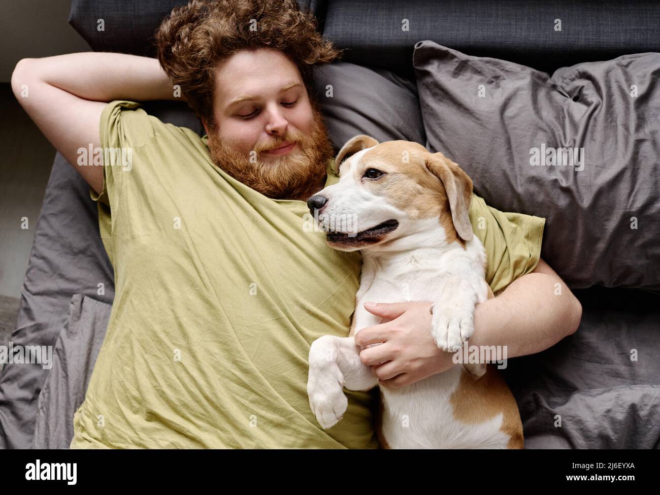High angle view of young bearded guy relaxing on bed in bedroom embracing his dog Stock Photo