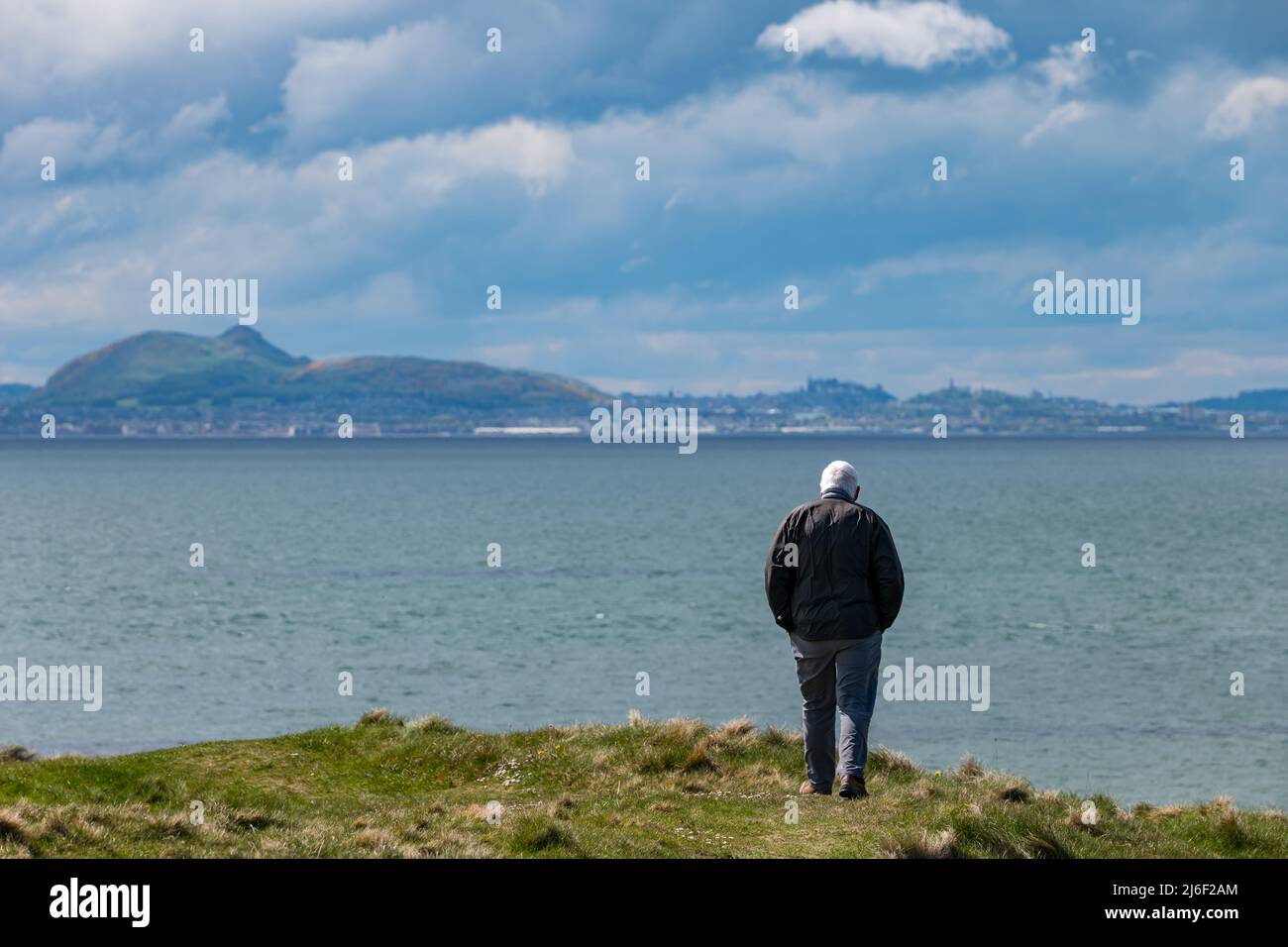 An older man walking on Firth of Forth coast with view of  Arthur's Seat and Edinburgh city skyline in the distance, East Lothian, Scotland, UK Stock Photo