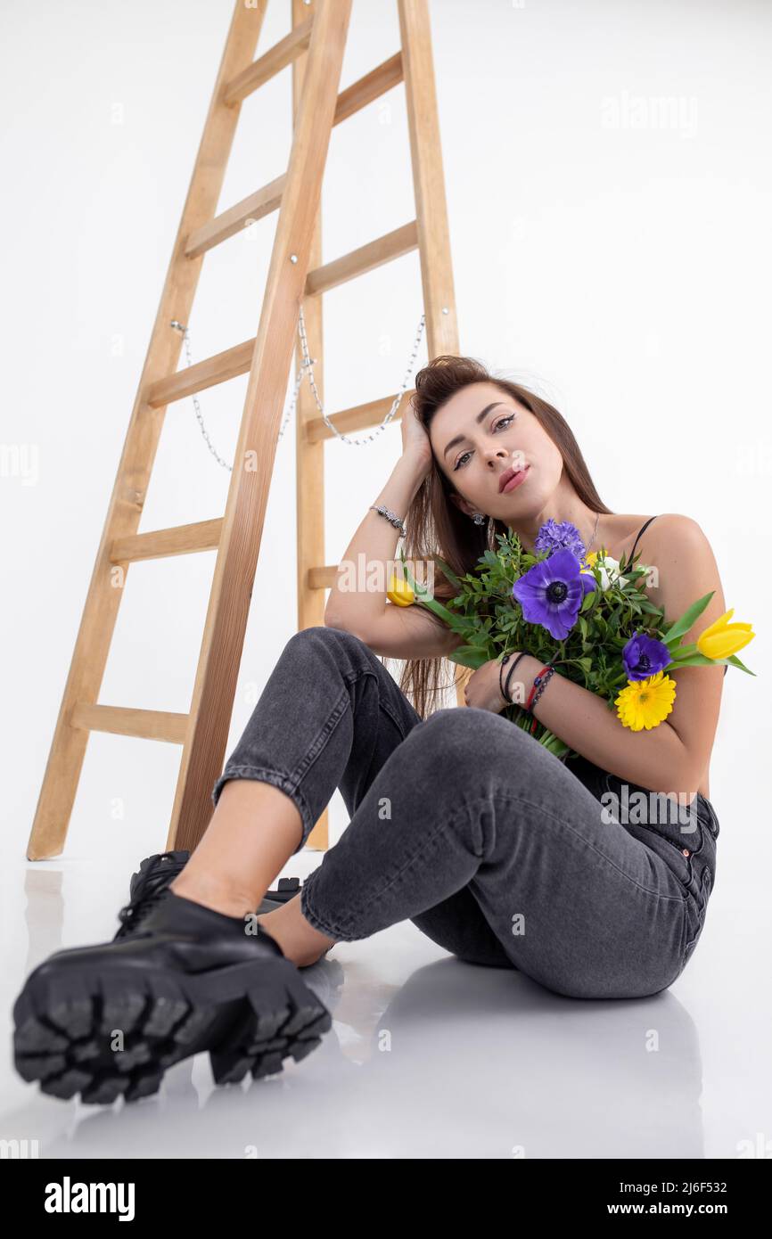 Portrait of young pretty woman with long dark hair wearing grey jeans, sitting near wooden ladder, holding bouquet. Stock Photo