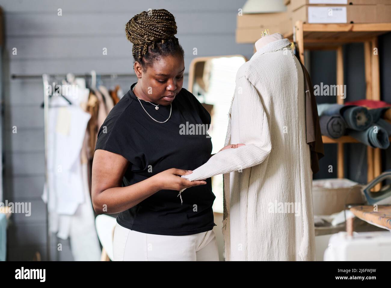 Young contemporary black woman in casualwear checking sewing quality of white coat on mannequin while looking at sleeve in workshop Stock Photo
