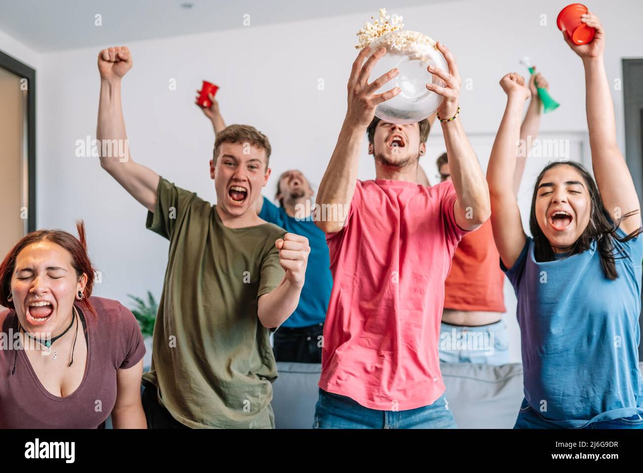 friends jumping for joy, throwing popcorn in the air after their team's victory. group of youngsters partying at home. Stock Photo