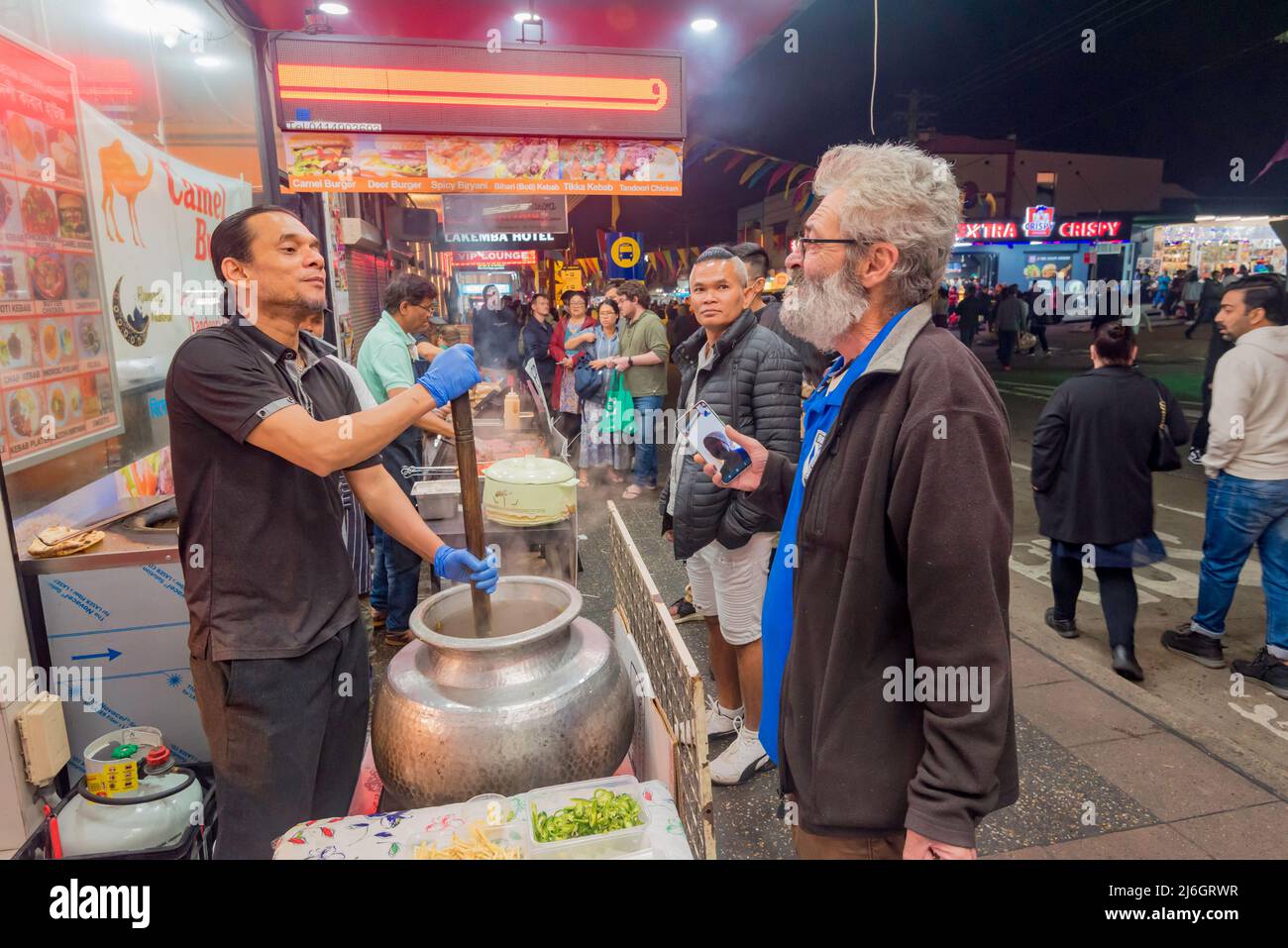 April 30, Lakemba, Sydney, Australia: On the final night of Ramadan in Sydney, the Ramadan Nights Festival is in full swing from sunset at 17.30 through to 02.00 the next morning. Here two men chat while one stirs a large pot of dal style food, possibly Dal Makhani, with a large wooden paddle. Stock Photo