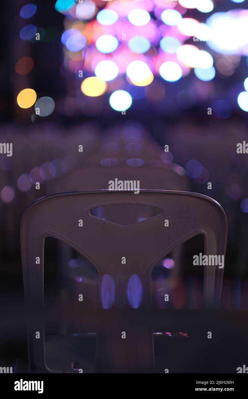 A strict row of empty plastic chairs, spaced according to social distancing, prepared for big event, in front of blurred colorful lights of a stage Stock Photo