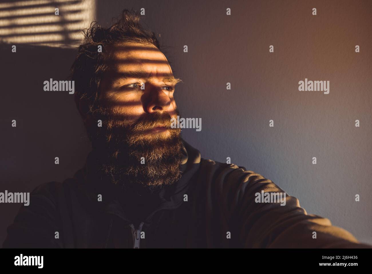 Hipster looking man hiding behind the venetian blinds or window shades. Cool guy wearng a hoodie in an interesting lighting. Stock Photo