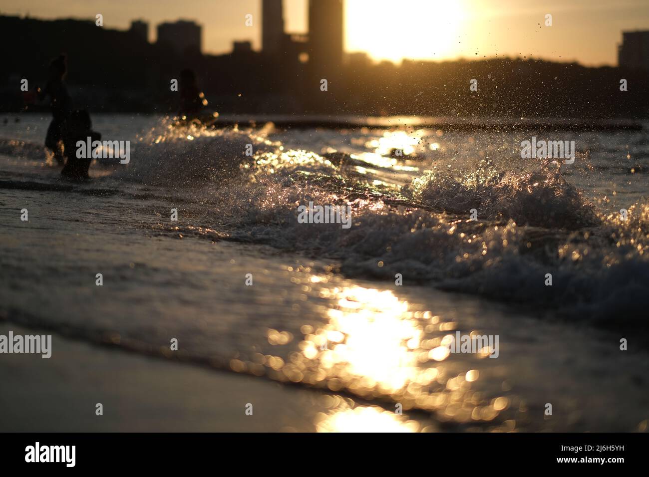 Waves are pounding on a sunlit beach, forming myriads of gold colored splashes, while people are having fun in a background Stock Photo