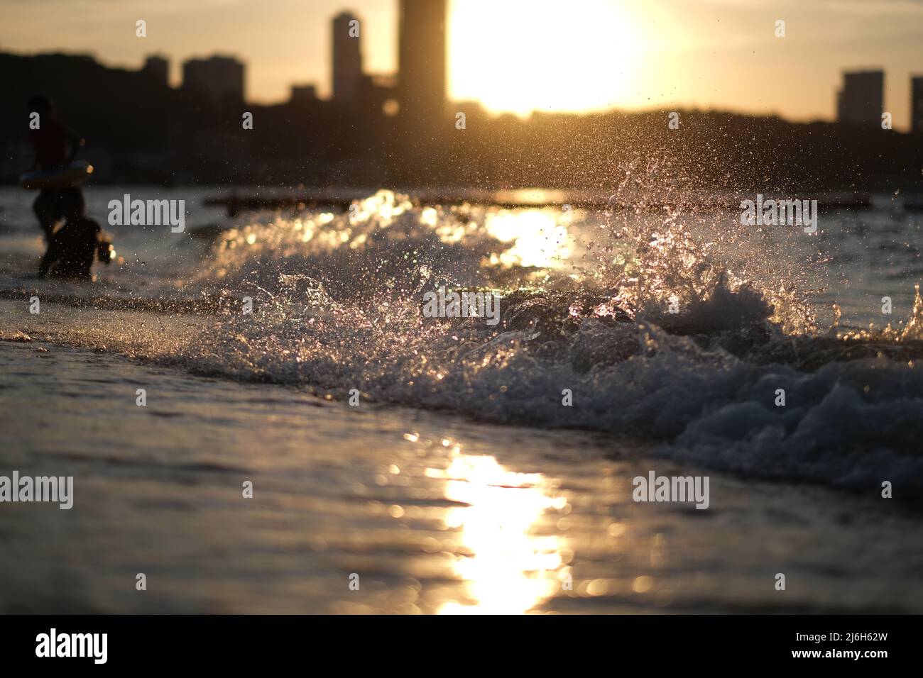 Waves are pounding on a sunlit beach, forming myriads of gold colored splashes, while people are having fun in a background Stock Photo