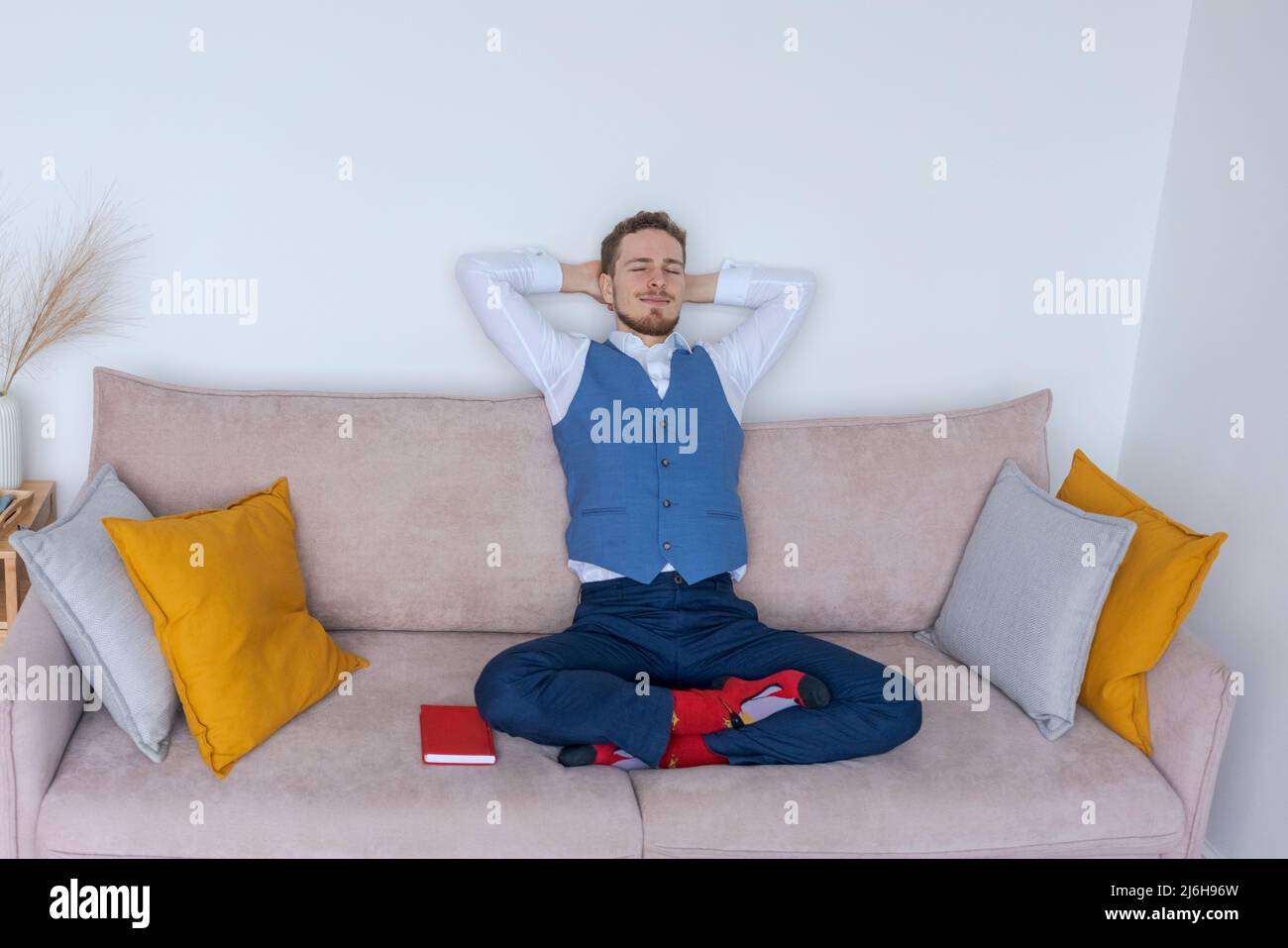 Successful young business man sits on sofa in lotus position in blue suit with notepad and thinks about business plans. Caucasian businessman meditates in the office on the couch Stock Photo