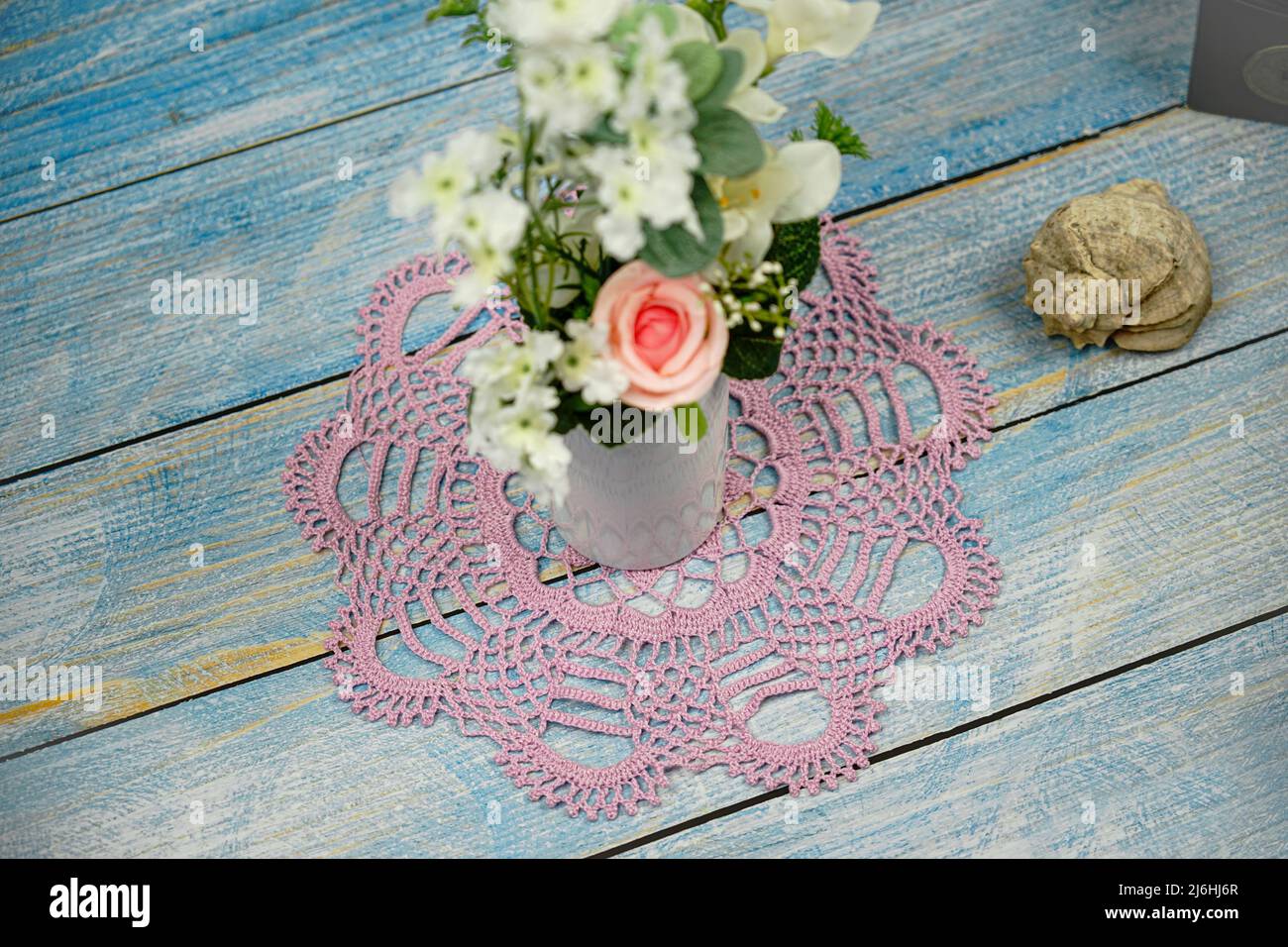 Hand made macrame lace napkin lies on a wooden windowsill. There is a vase of flowers on a lace Stock Photo
