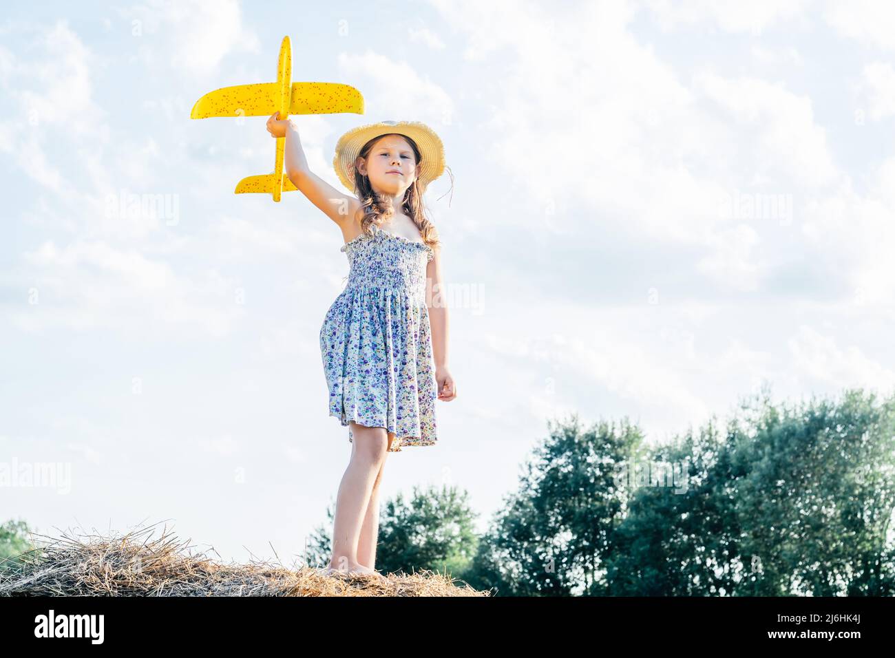 Portrait of little girl in hat and dress playing flying yellow toy airplane, standing on haystack. Taking aim in sky. Light sunny day. Having Stock Photo