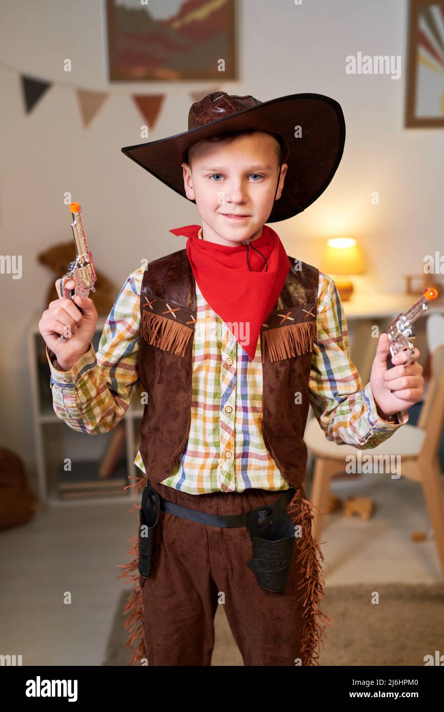 Happy cute little boy in costume of cowboy posing for commercial with two revolvers while standing in his room and looking at camera Stock Photo