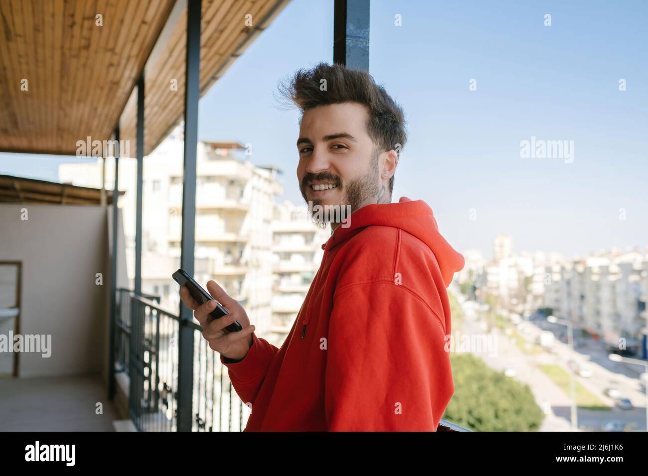 Looking at the camera, young man leaning on balcony rail and posing for camera, handsome adult holding phone surfing internet and hanging out on socia Stock Photo