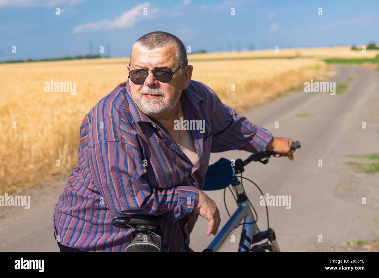 Outdoor portrait of a bearded, chubby Ukrainian senior man leant elbows to handle bar of his bicycle when doing  short rest on summer ride on a countr Stock Photo