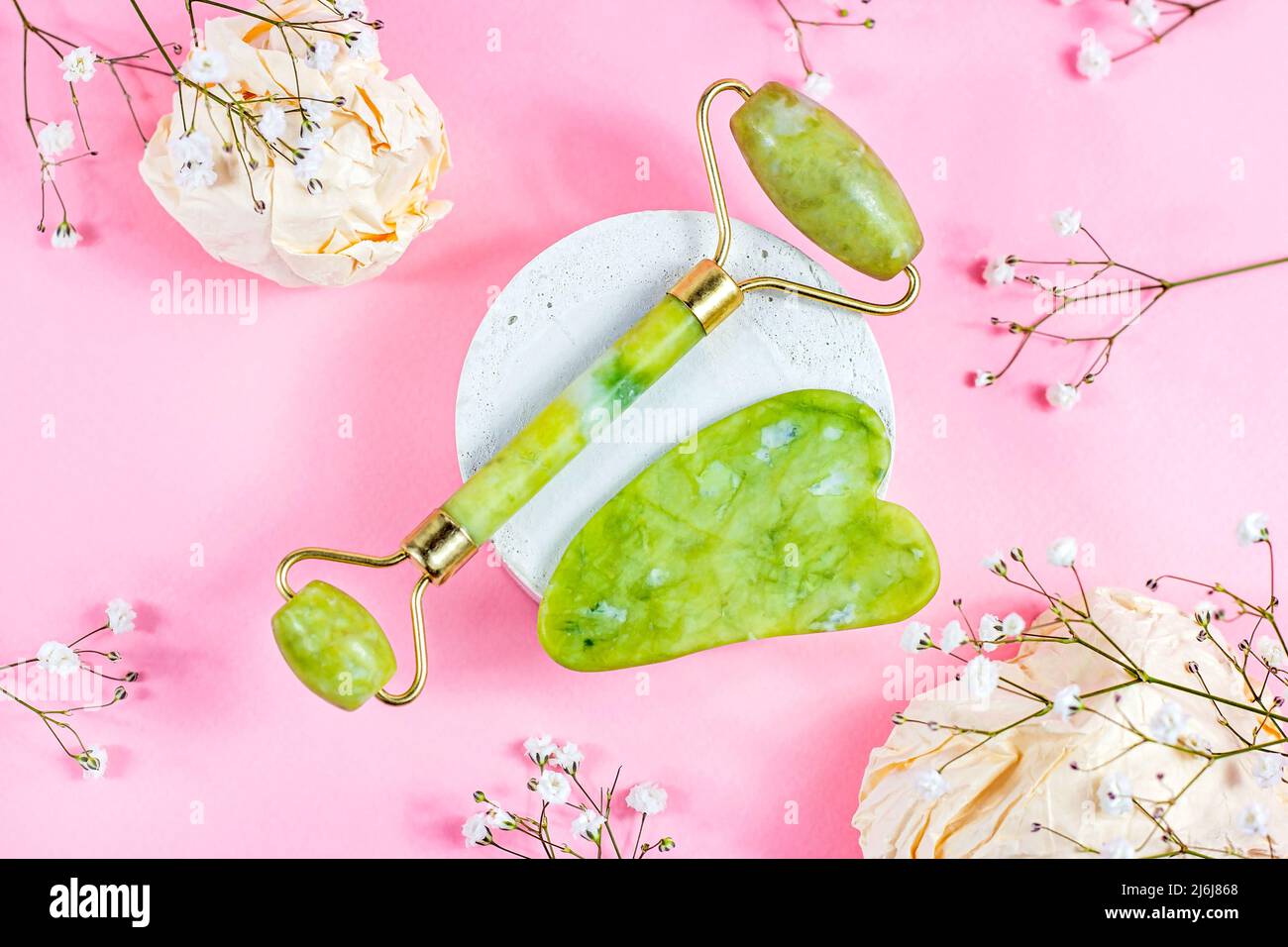 Green quartz stone gua sha scraper and jade roller for facial beauty lifting anti age massage with white flowers on pastel pink background top view. Stock Photo