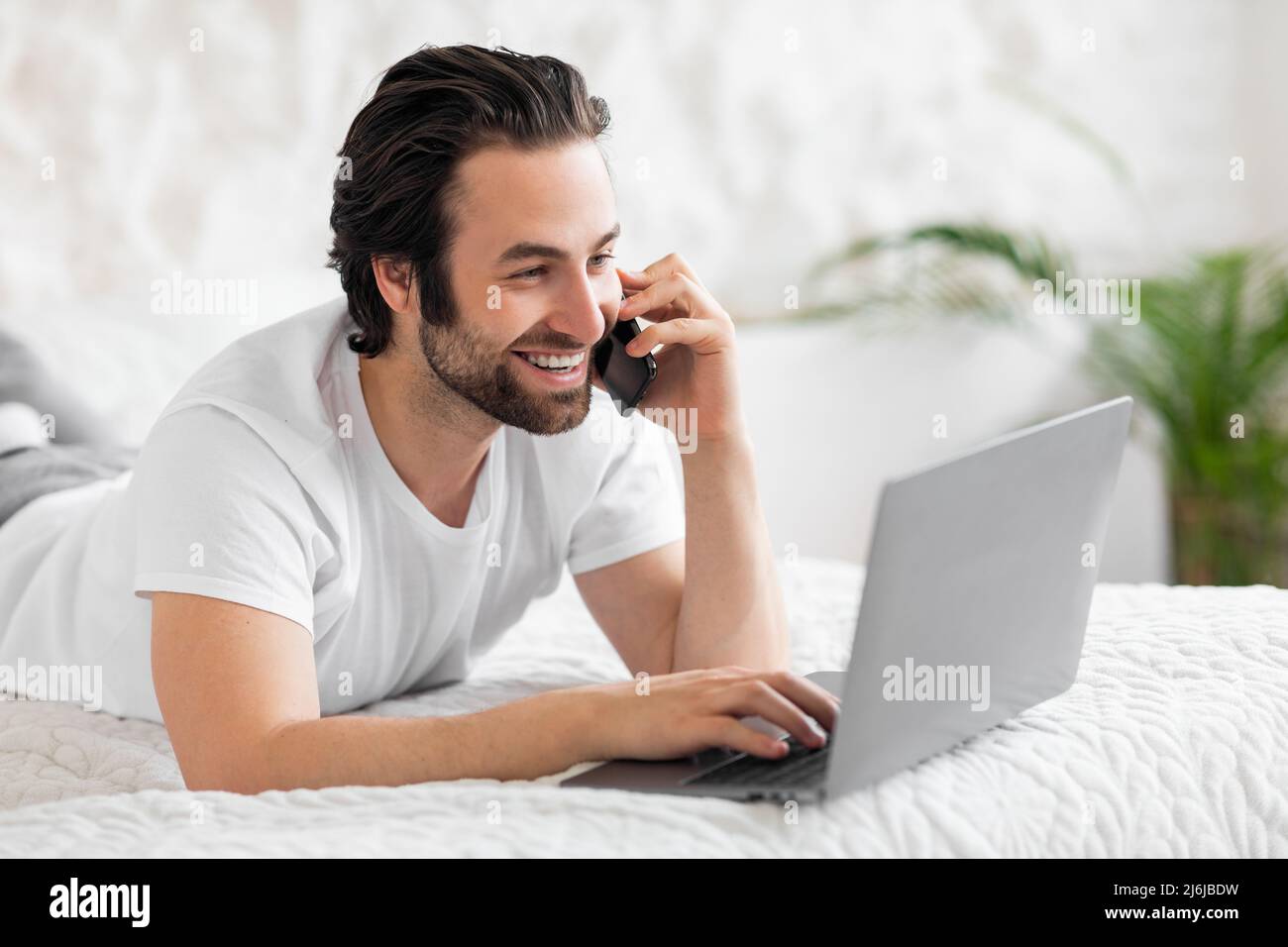 Smiling man making phone call and using laptop in bed Stock Photo