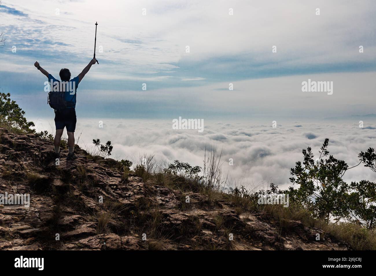 Hiker Asian man in silhouette standing on peak of mountain with cloud scape raises hand to signify success Stock Photo