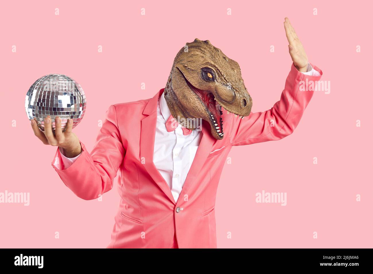 Fashionable man in dinosaur rubber mask with disco ball in his hand on pastel pink background. Stock Photo