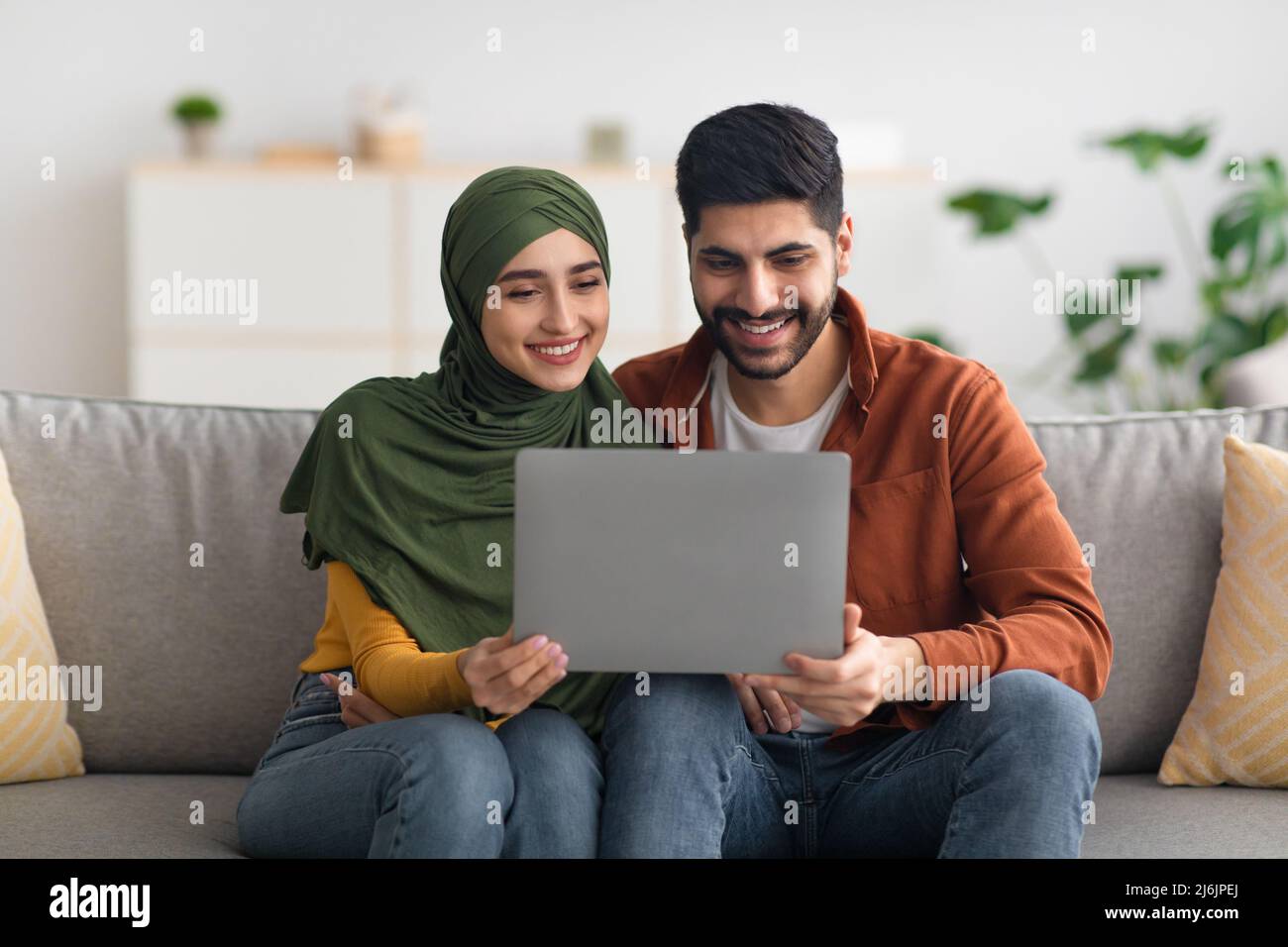 Happy Middle Eastern Spouses Using Laptop Sitting On Couch Indoor Stock Photo