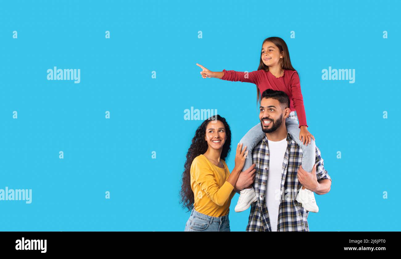 Arabic Girl Pointing Finger Sitting On Father's Shoulders, Blue Background Stock Photo