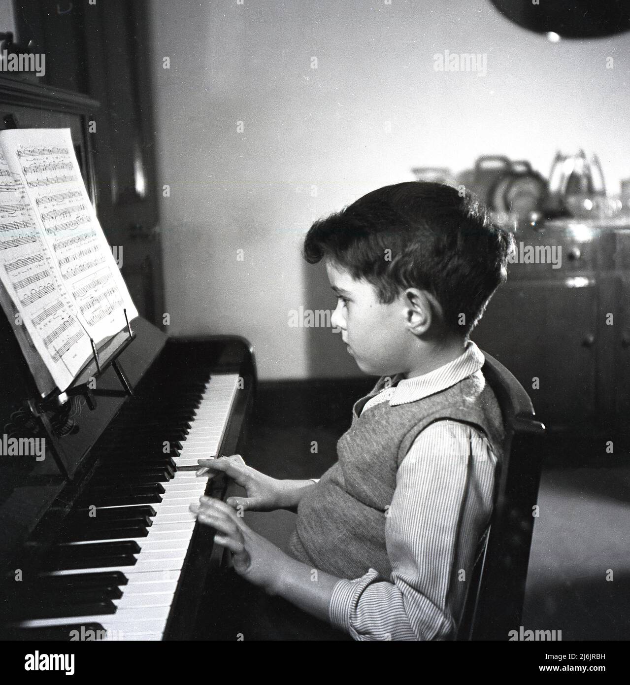1953, historical, inside a room at home, a young boy sitting playing a traditional upright piano, with sheet music above the keyboard, England, UK. Stock Photo
