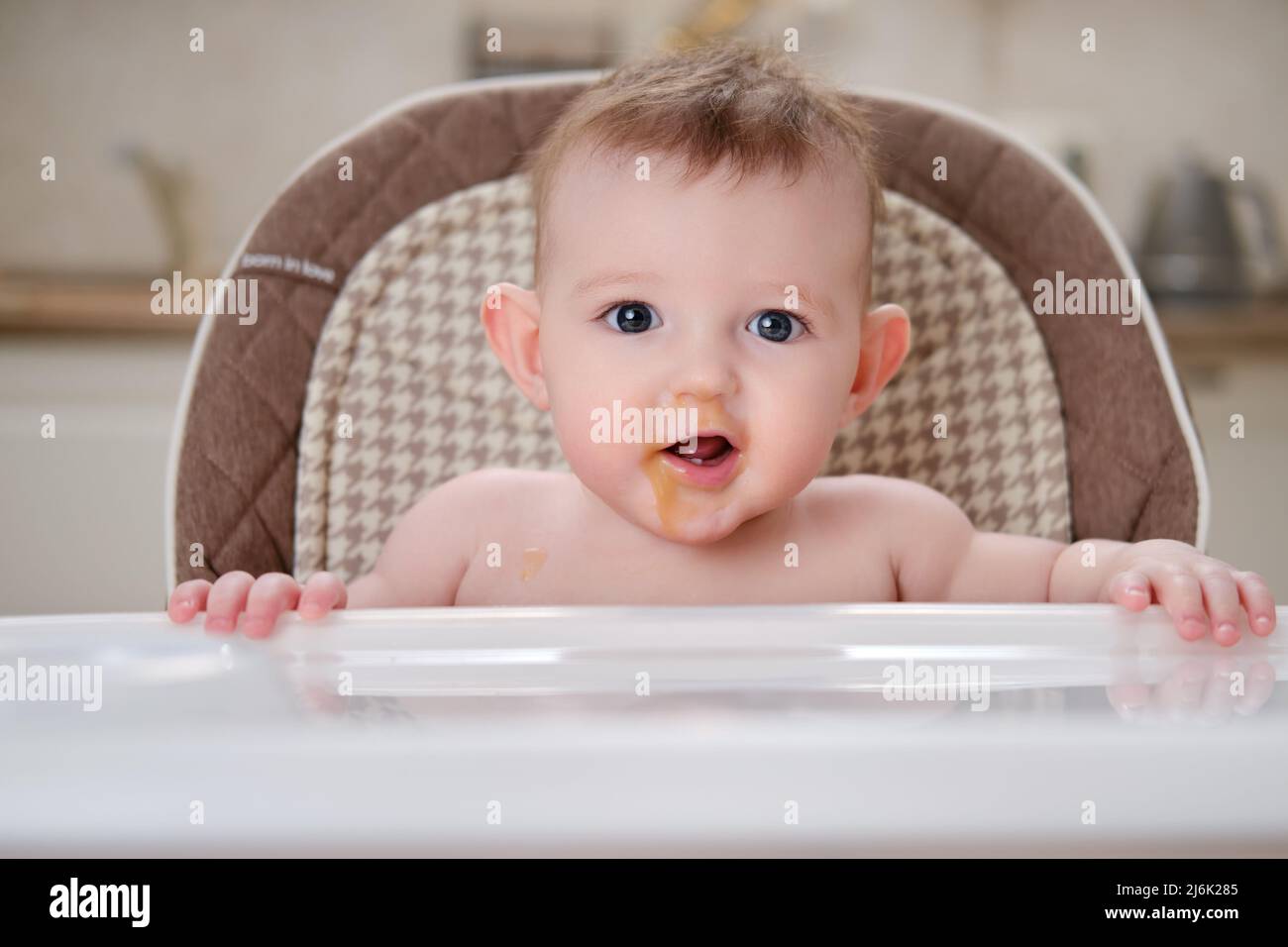 Happy baby with food stained mouth sitting on child chair, home kitchen background Stock Photo