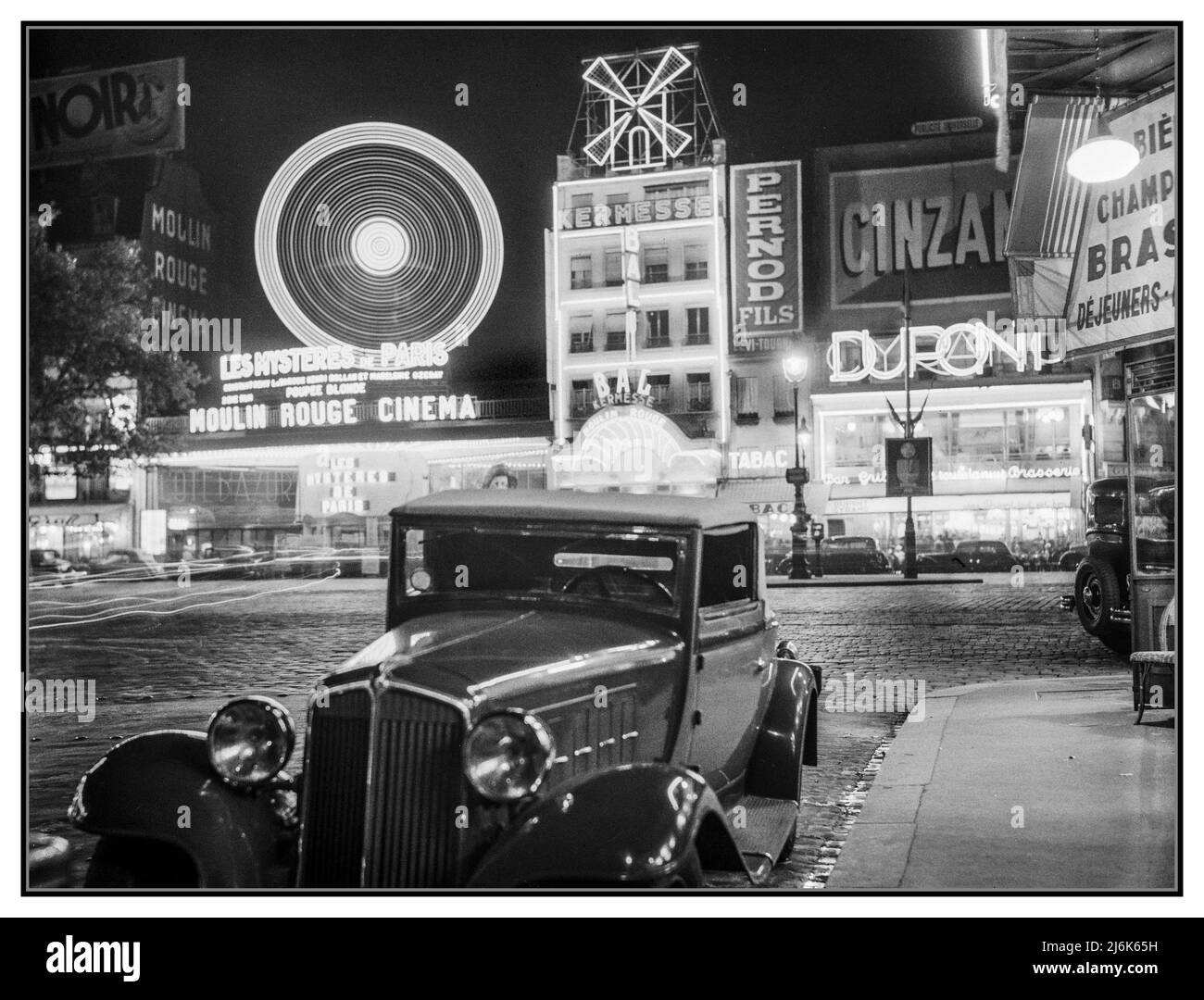 MOULIN ROUGE Retro Vintage Paris night lights advertising neon B&W The Moulin Rouge Montmartre Paris France at night 1936: 2 door coupe car in foreground, French drinks alcohol advertising hoardings France, Paris Moulin Rouge Photographer :  Willem van de Poll Stock Photo