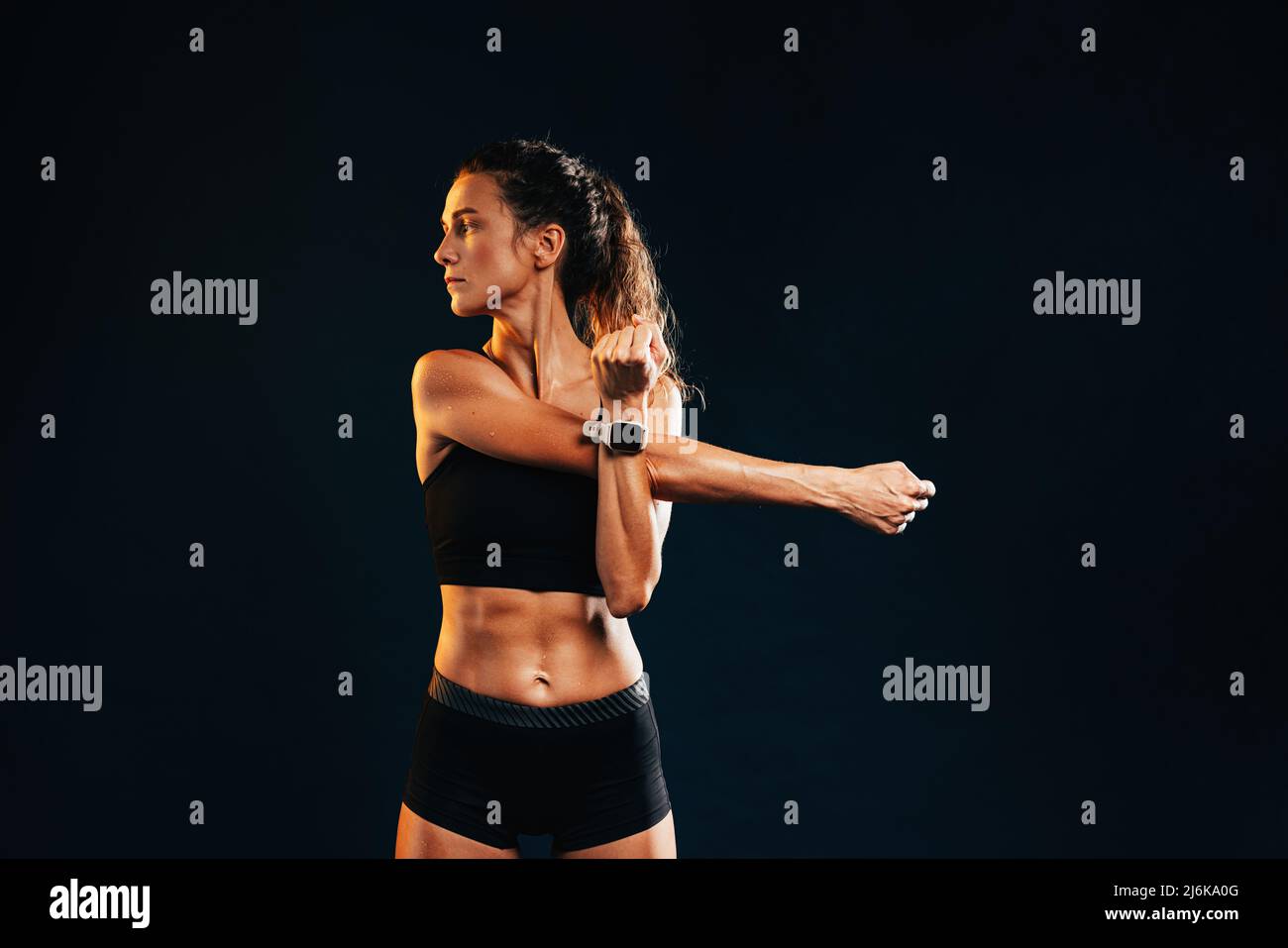 Young muscular woman stretching her arm in studio on black background looking away Stock Photo