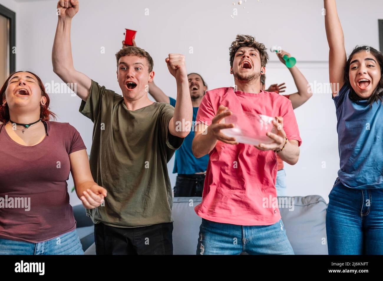 friends jumping for joy, throwing popcorn in the air after their team's victory. group of youngsters partying at home. Stock Photo