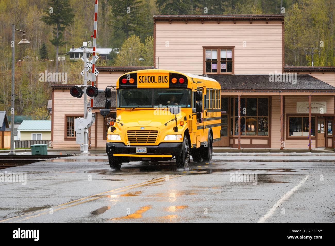 Skykomish, WA, USA - April 26, 2022; Yellow school bus crossing railway tracks in Skykomish Washington.  The road is wet in the rain. Stock Photo