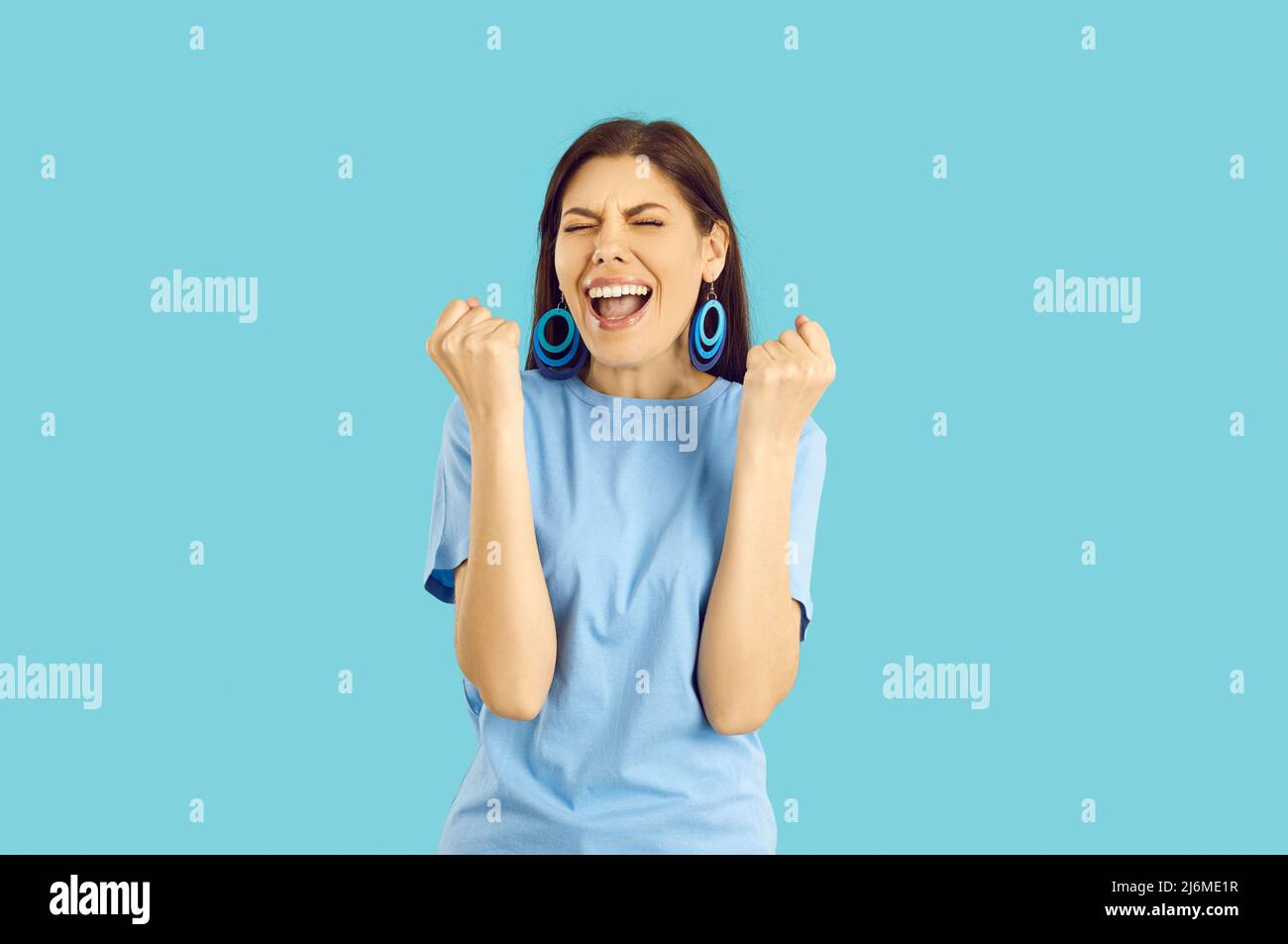 Happy excited young woman standing on blue background and screaming YES and HOORAY Stock Photo