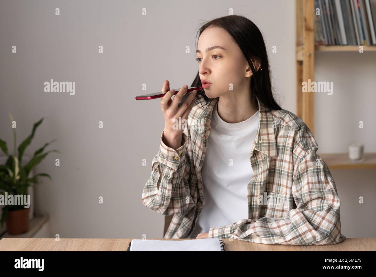 Beautiful caucasian teenage girl giving voice commands to virtual assistant on smartphone, talking while recording audio messages, using mobile phone Stock Photo