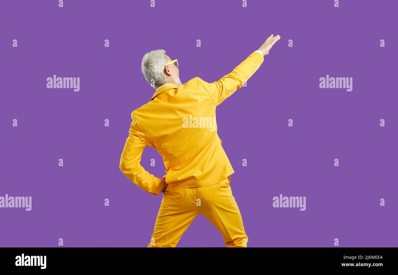 Back view of funny senior guy in yellow party suit dancing isolated on purple background Stock Photo