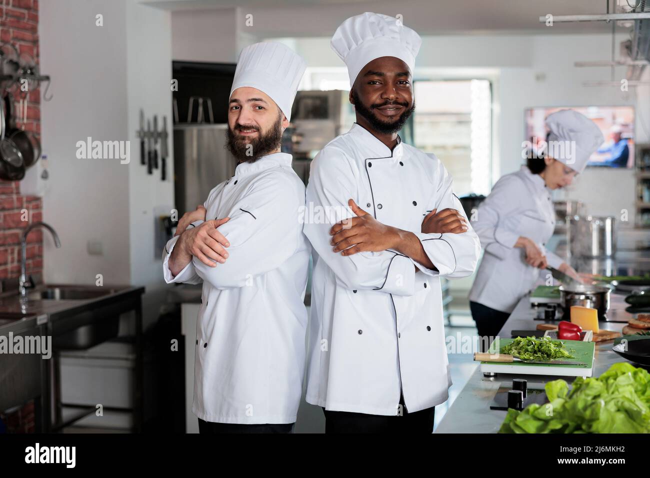 Confident smiling chefs standing in restaurant kitchen with arms crossed while preparing