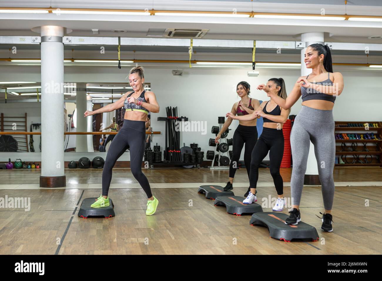 Group of gleeful female athletes and instructor smiling and dancing energetically on steppers during aerobic training in spacious modern gym. Stock Photo