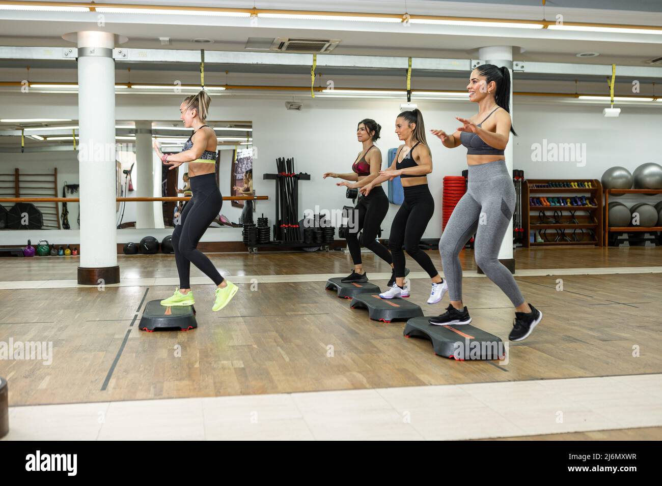 Group of female athletes and trainer smiling and doing exercise on steppers during aerobic workout in light gym. Stock Photo
