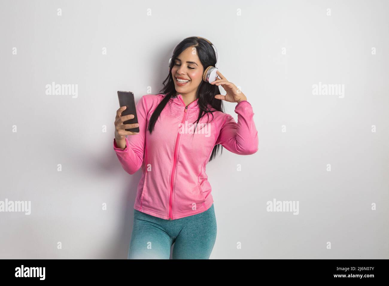 Cheerful Hispanic sportswoman in activewear searching playlist for training on cellphone while enjoying music in headphones in studio against white Stock Photo