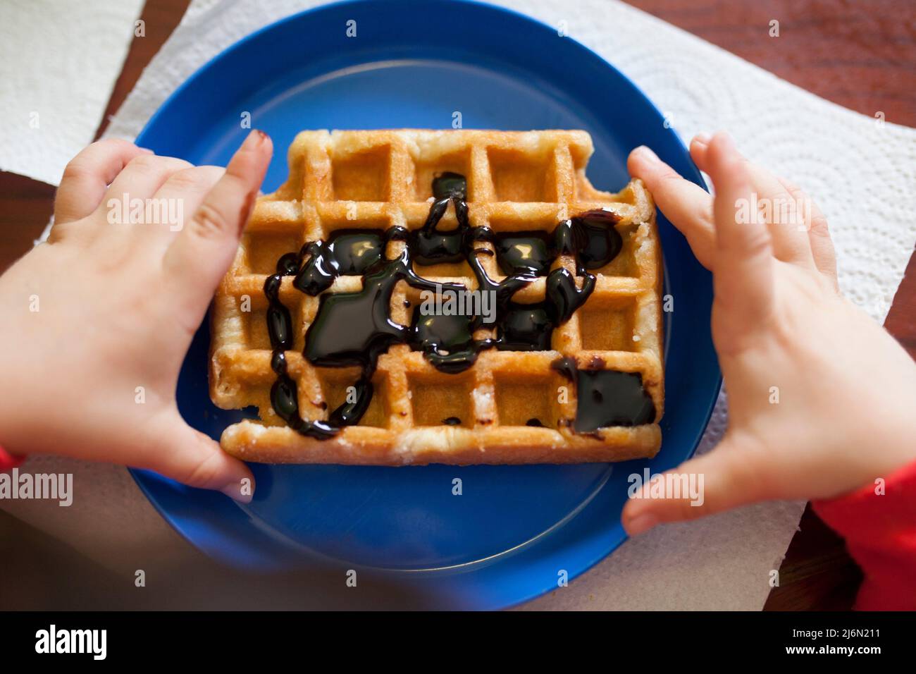 Child holds a waffle over blue plate. Waffled is filled with chocolate syrup. Stock Photo