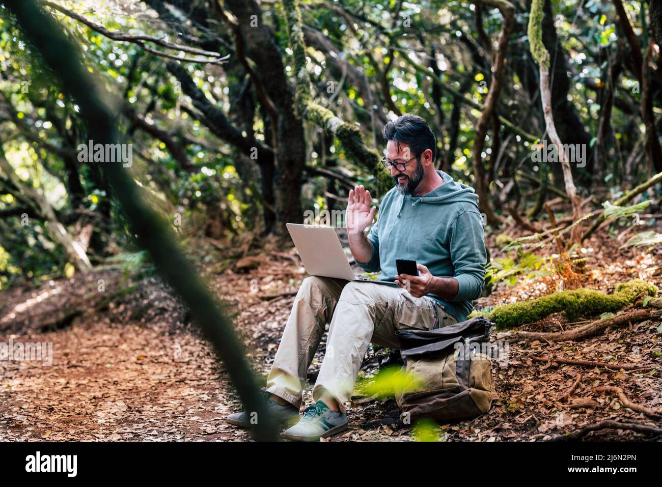 Adult caucasian man sitting on the ground in the forest do a video call connection with laptop computer - internet wireless everywhere and phone Stock Photo