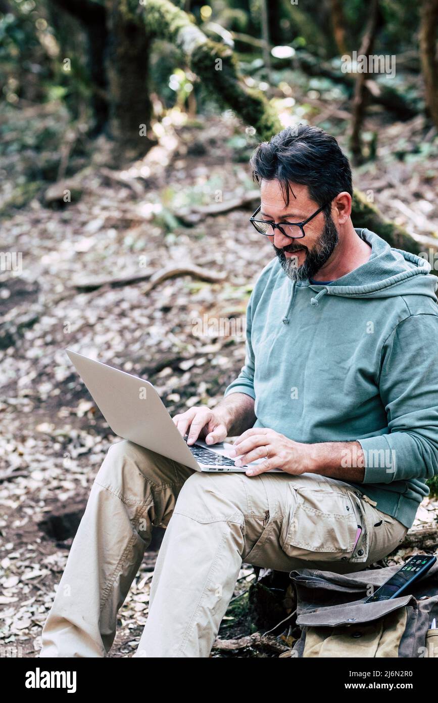 Adult caucasian man sitting on the ground in the forest do a online work connection with laptop computer - internet wireless smart working job Stock Photo