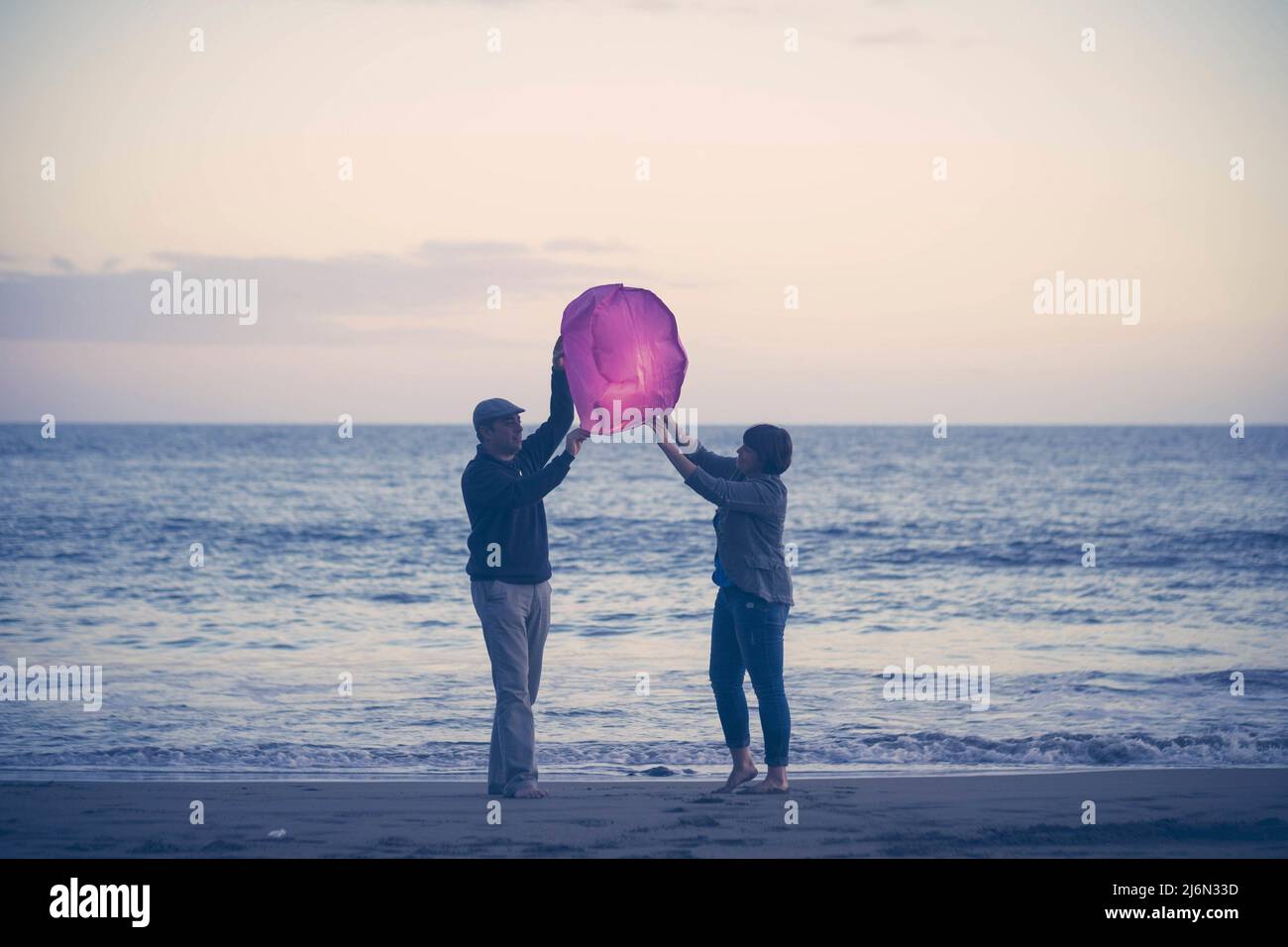 Couple on beach flying paper lantern. Middle aged couple with chinese lantern during sunset at beach. Joyful couple about to release paper lantern in Stock Photo