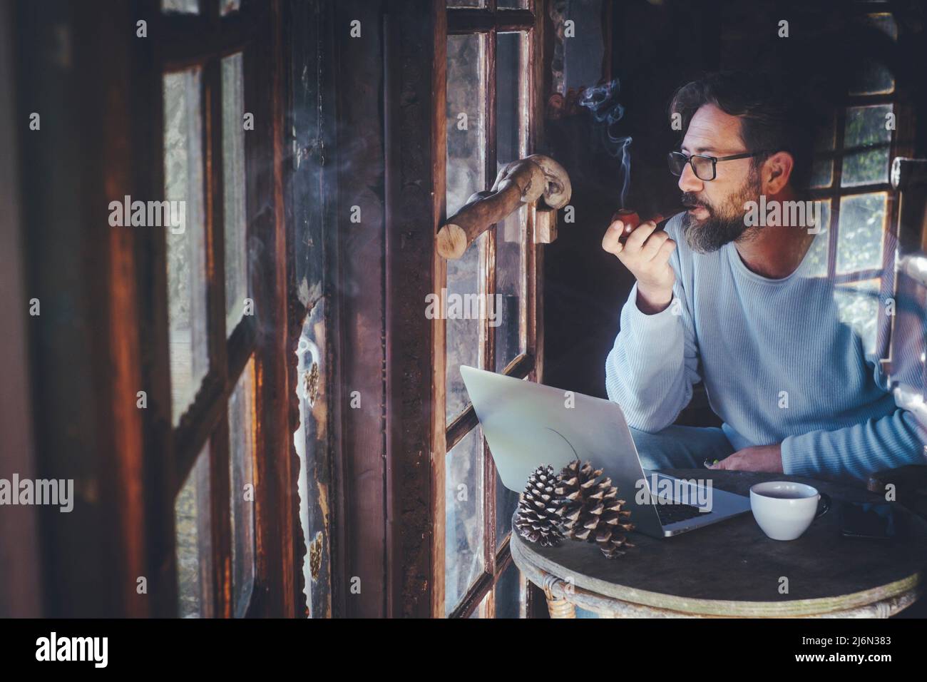 Mature hipster man use computer laptop at home in chalet wooden house smoking pipe and tobacco alone. Senior modern people lifestyle with connection Stock Photo