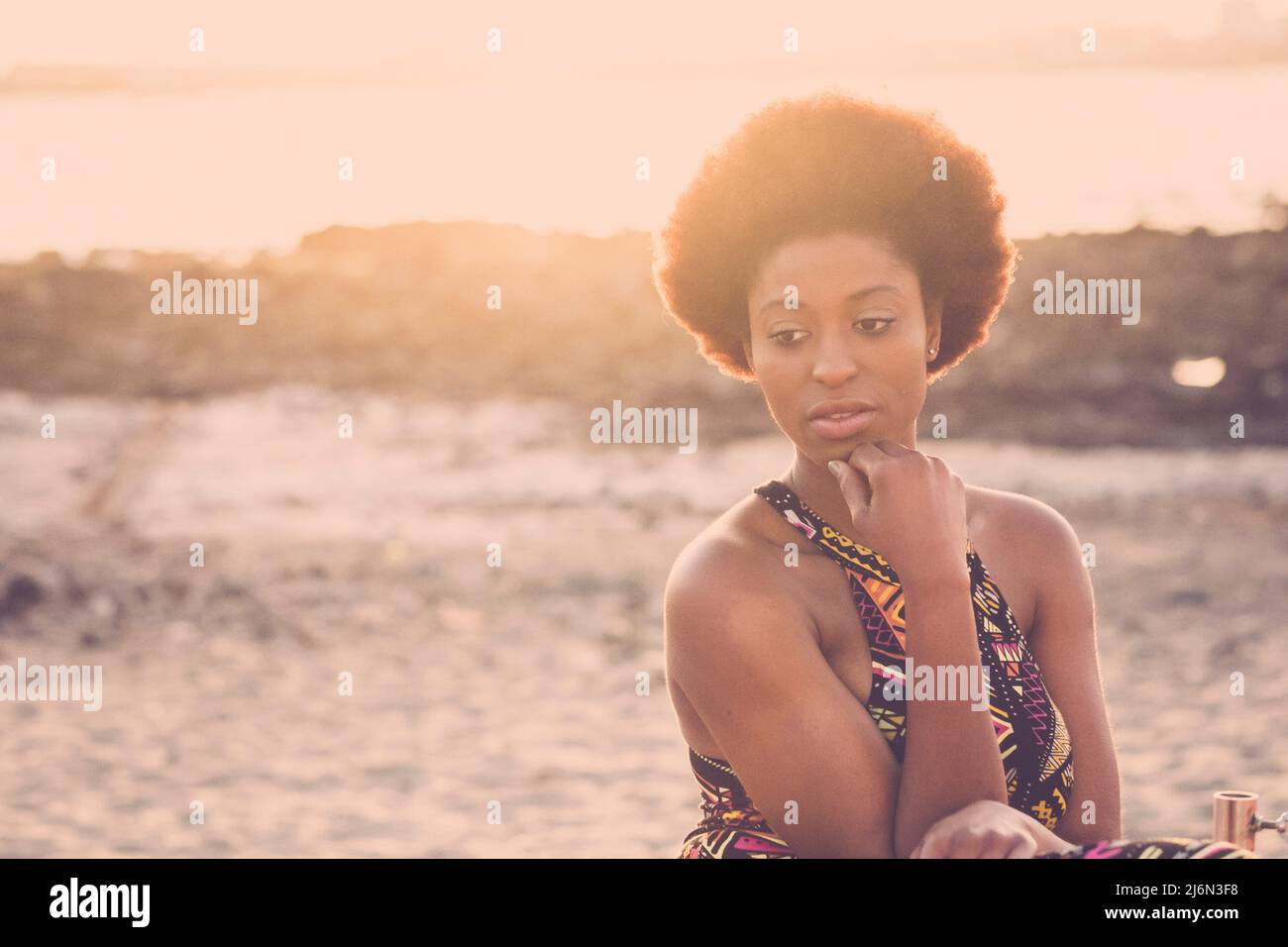 Thoughtful african american woman sitting on beach with hand on chin. Attractive black woman relaxing on sandy beach during holidays. Young woman Stock Photo